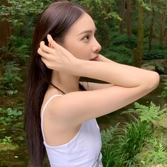 a woman with long brown hair standing in front of a river and trees wearing a white tank top