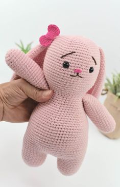 a hand holding a pink crocheted stuffed animal in front of a potted plant