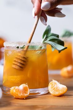 a hand holding a wooden stick over a glass filled with orange juice and garnished with leaves