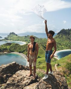 two shirtless men standing on top of a mountain next to each other holding a white kite