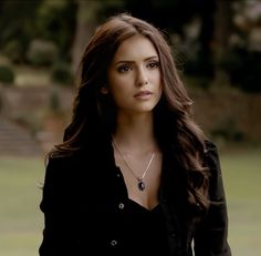 a woman with long brown hair wearing a black shirt and silver necklace standing in front of a grassy area