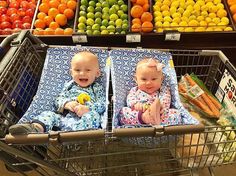 two babies in pajamas sitting in a shopping cart at a grocery store with fruits and vegetables behind them