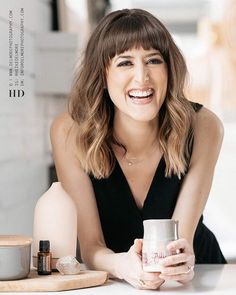 a woman sitting at a table holding a coffee cup and smiling for the camera with her hands