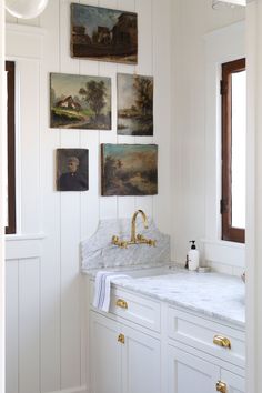 a white bathroom with gold faucet and marble counter top, paintings on the wall