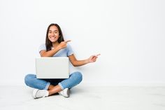 a young woman sitting on the floor with her laptop pointing to the side and smiling