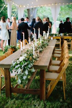 a long wooden table topped with candles and greenery next to a white tented area