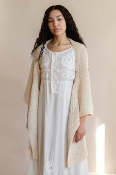 a woman standing in front of a wall wearing a white dress and cardigan sweater