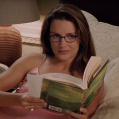 a woman laying in bed reading a book with glasses on her face and looking at the camera