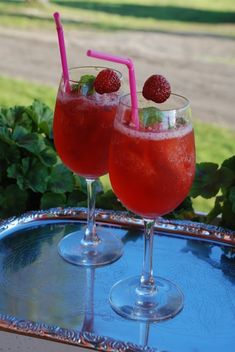 two glasses filled with liquid and strawberries on top of a metal tray next to bushes