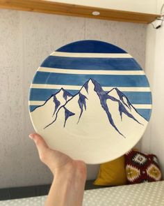 a hand holding up a plate with mountains painted on the front and sides, in front of a wall