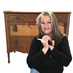 a woman sitting in front of a wooden dresser holding a pencil and looking at the camera