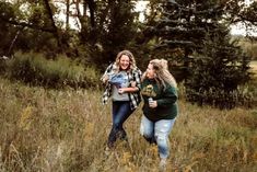 two women walking through tall grass in the woods, one carrying a baby and smiling at the camera