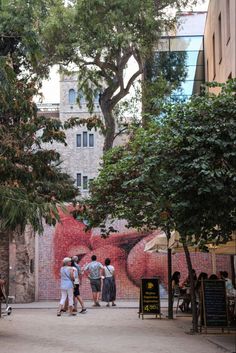 some people are walking down the street near many trees and buildings with graffiti on it