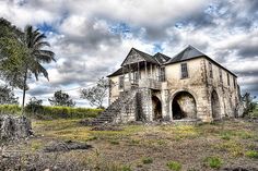 an old abandoned house sits in the middle of a field with palm trees and cloudy skies