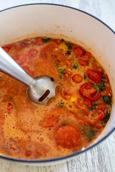 a pot filled with tomato soup on top of a white wooden table next to a spoon