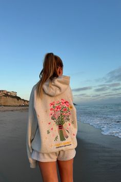 photo shows a blonde teenage girl standing on a sandy beach at sunset. she is facing away from the camera and is looking at the ocean. she is wearing gray sweat shorts and a gray hoodie that has pink embroidered flowers on the back and the words "gift giving" and "courtesy of dandy worldwide." the ocean is to the right of the girl and the sandy beach is to the left. Dandy Hoodie Gift Giving, Dandy World Wide Hoodie, Dandy Hoodie Outfit, Dandy Sweatshirt, Dandy Hoodie, Madhappy Hoodie, Hoodies Design Ideas