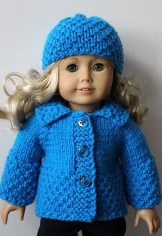 a doll with blonde hair wearing a blue knitted jacket and hat, standing next to a white wall