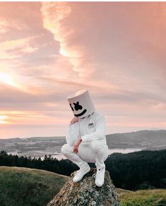 a man in a white suit and hat sitting on top of a rock with the sun setting behind him