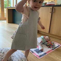 a young child standing on top of a wooden floor next to a baby doll in a kitchen