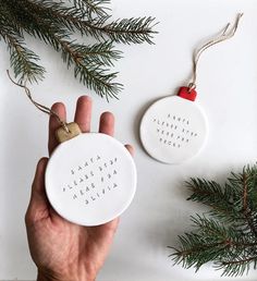 two white ceramic ornaments with words on them and pine branches in front of the ornament