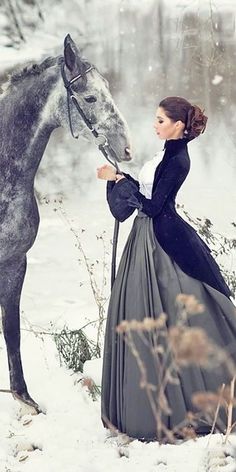 a woman in a long dress standing next to a horse on snow covered ground with trees and bushes behind her