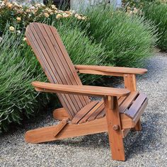 a wooden lawn chair sitting in front of some bushes and flowers on a gravel ground