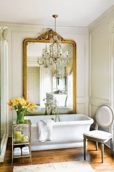 a white bath tub sitting under a large mirror next to a table with flowers on it