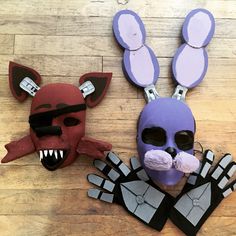 two paper mache masks on a wooden floor, one is purple and the other is black