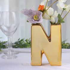 a gold letter shaped vase with flowers in it sitting on a table next to wine glasses