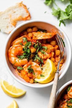 two bowls filled with shrimp and chickpeas next to lemon wedges on a white surface