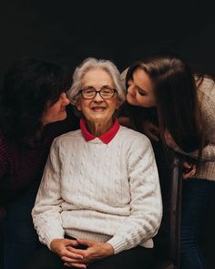 an older woman sitting in a chair with two younger women and one is kissing her