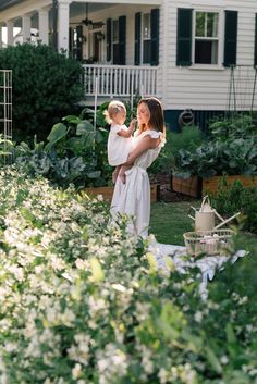 a woman holding a baby in her arms while standing next to a house and garden