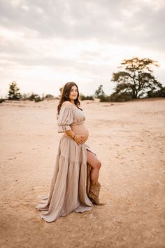 a pregnant woman sitting in the sand with her legs crossed and wearing cowboy boots, posing for a photo