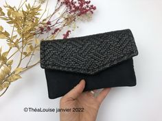 a hand holding a black clutch bag next to some dried plants and flowers on a white surface