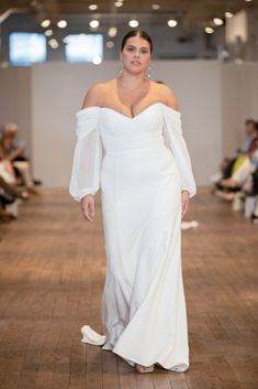 a woman walking down a runway wearing a white dress with long sleeves and an off the shoulder top