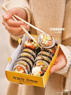 a person holding a yellow box with sushi in it and chopsticks sticking out