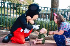 a woman kneeling down next to a baby near a mickey mouse statue and a small child