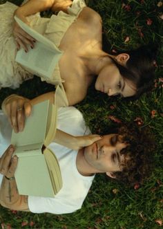 two people laying in the grass with books on their laps and one person holding an open book