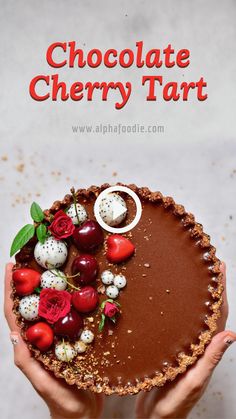 someone holding a chocolate tart with cherries and flowers on the top, in front of a white background