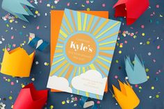 colorful paper crowns and confetti are scattered around the birthday greeting card that says kyle's first birthday