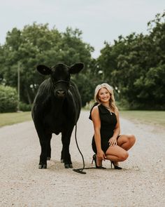 a woman kneeling down next to a black cow on a dirt road with trees in the background