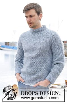 a man standing in front of a body of water wearing a blue sweater and jeans