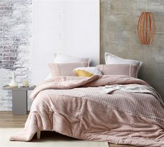 a bed with pink comforter and pillows in a white room next to a brick wall