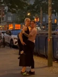 two people are standing on the sidewalk in front of some parked cars and one is hugging another