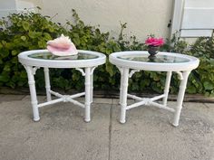 two white side tables with glass tops and flowers on each end table, sitting in front of some bushes