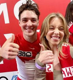 two people giving the thumbs up sign in front of a red and white wall with words on it