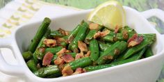 a white bowl filled with green beans and bacon next to a slice of lemon wedge