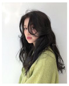 a woman with long black hair wearing a green sweater and looking at the camera while standing against a white wall