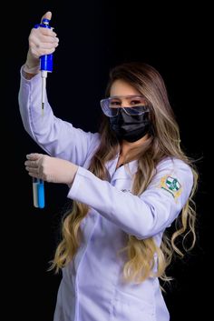 a woman in white lab coat and goggles holding up a blue toothbrush wearing a black mask