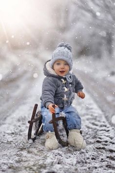 a baby boy sitting on top of a snow covered ground holding an umbrella and wearing a hat